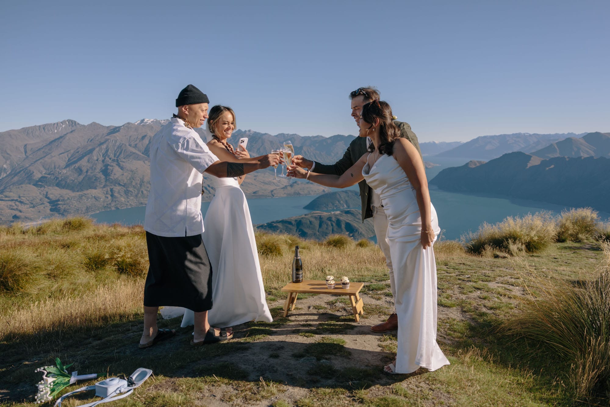 A toast on the mountainside above the lake, glasses raised, with cake and a bottle on a low wooden table