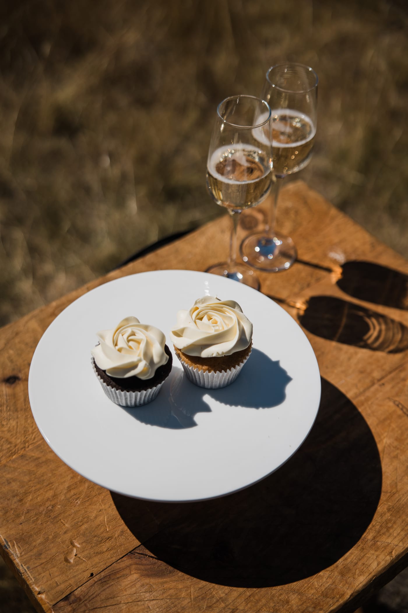 Two cupcakes iced with white roses on a plate, and two filled glasses, on a rough wooden table in the dry grass