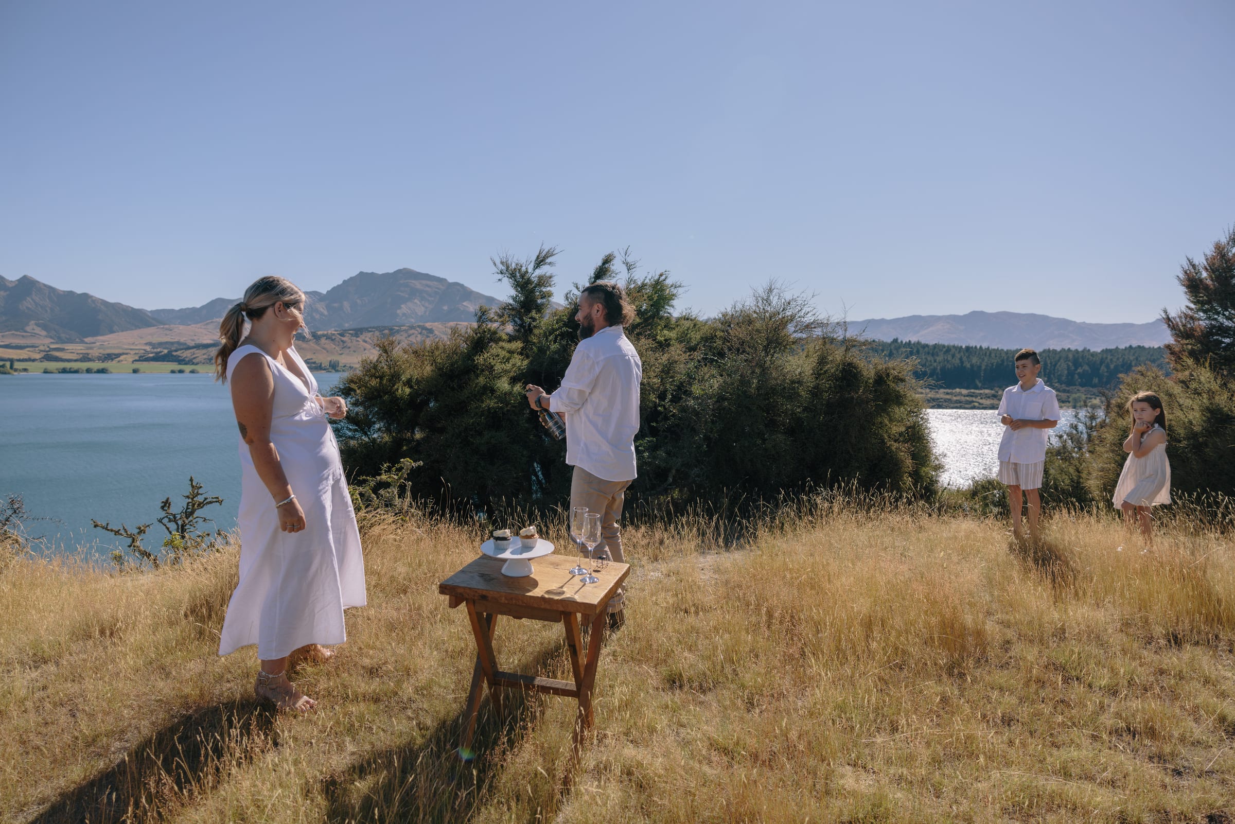 A small group on a tussock hillside above the lake, someone pouring at a low wooden table set with cups, two children waiting to one side